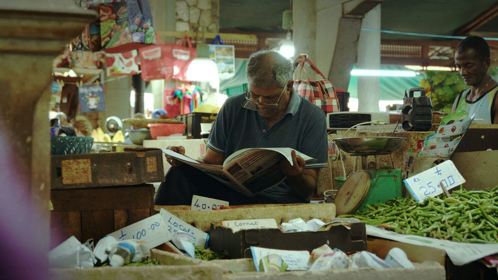 A man reading behind a vegetable stall in a Mauritian market, from Groundwork